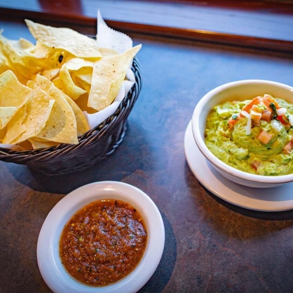 A basket of tortilla chips, a bowl of guacamole, and a bowl of salsa on a dark table.