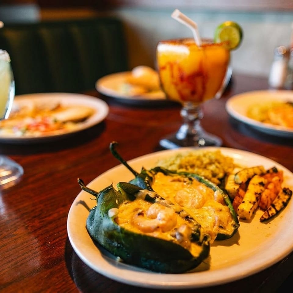 Two shrimp-stuffed poblano peppers, rice, and grilled vegetables with two margaritas on a wooden table.