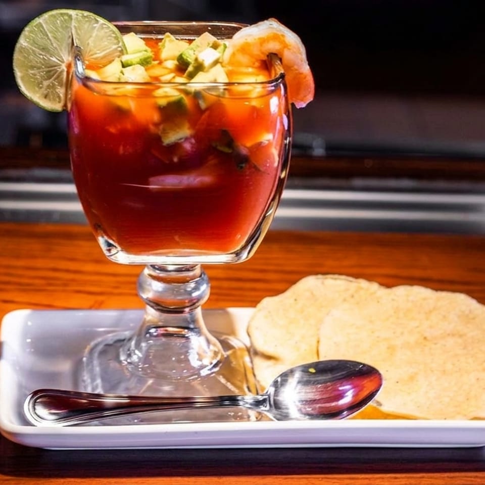 Shrimp cocktail with avocado, lime, and a side of tostadas and a spoon on a wooden table.