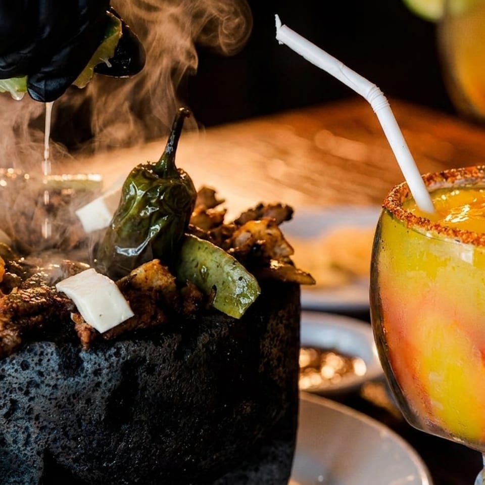 Steaming molcajete with meat, shrimp, cheese, and peppers, next to a mango margarita.